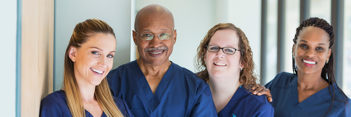 A team of four multi-ethnic medical professionals standing in a corridor, wearing blue scrubs, smiling